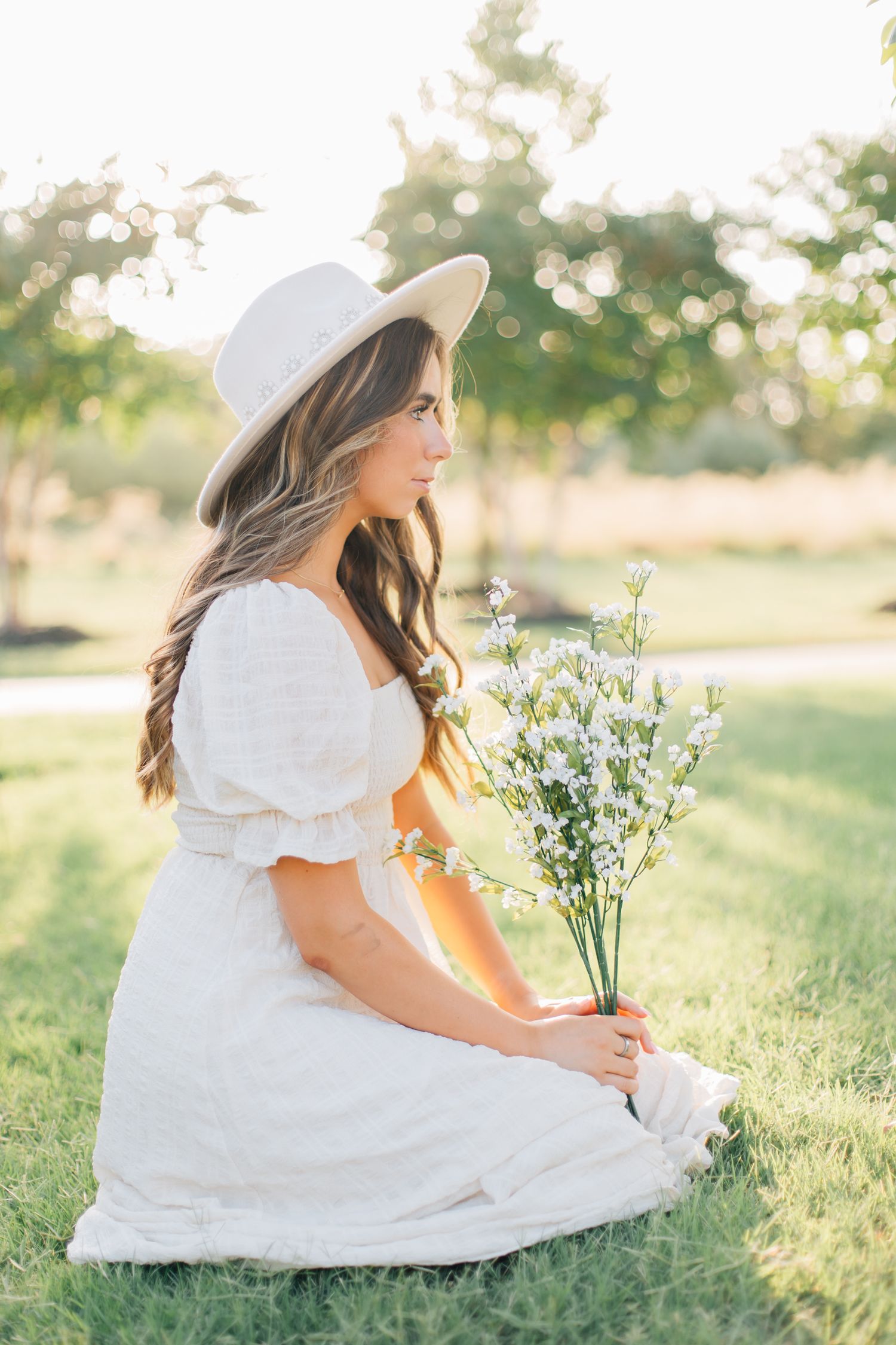 Woman in white dress and sun hat holds wildflowers while sitting in a sunny meadow.