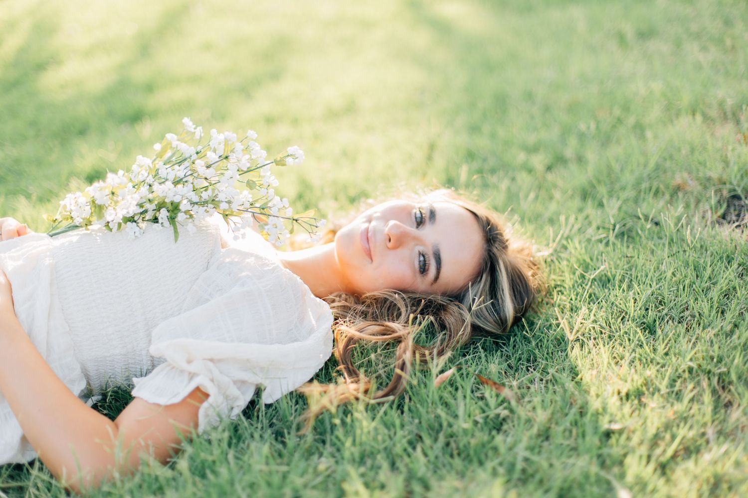 Woman in white dress lying on grass with wildflowers in dreamy summer portrait.