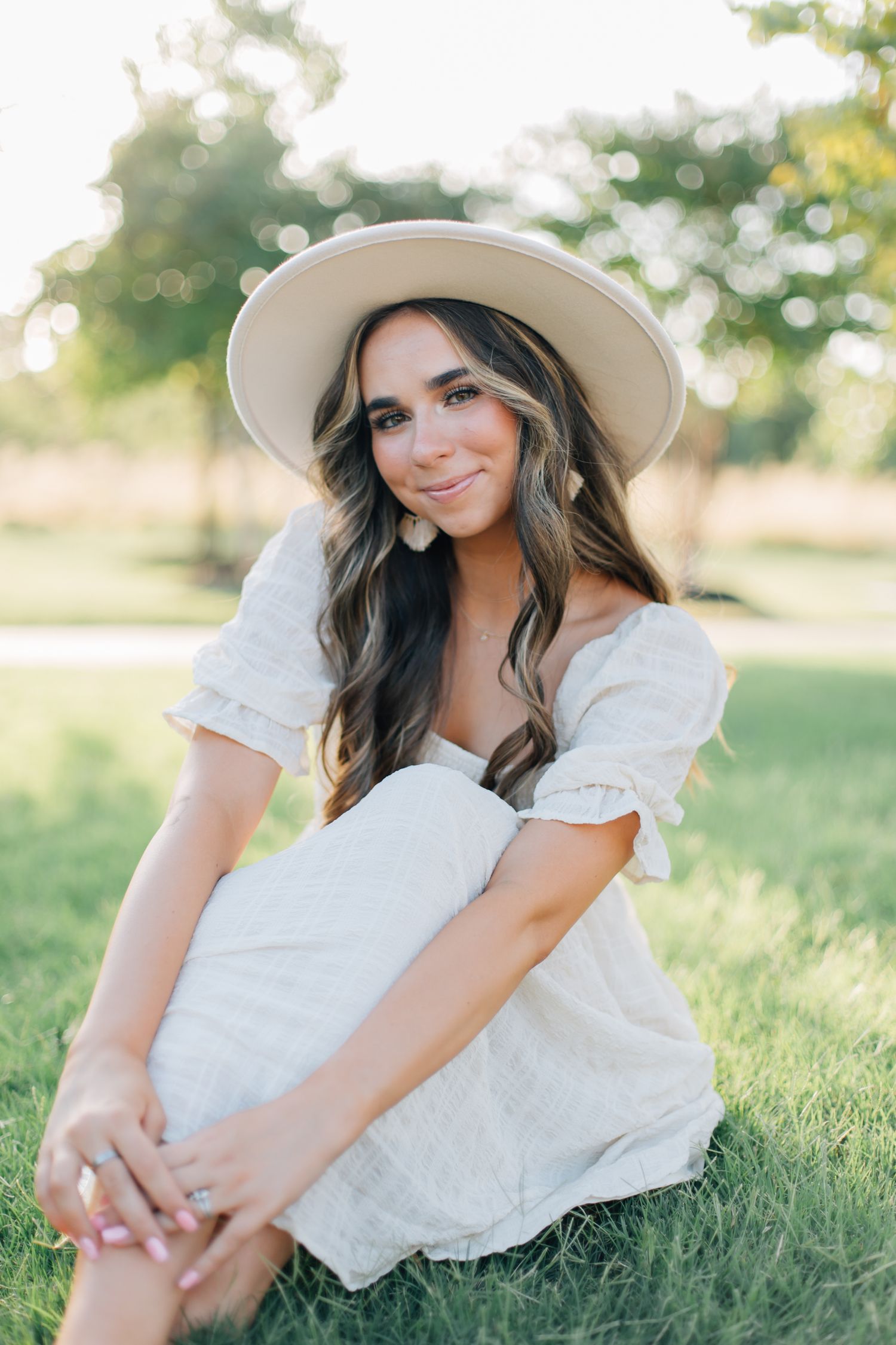 Woman in white dress and sun hat sitting on grass during golden hour photo session.