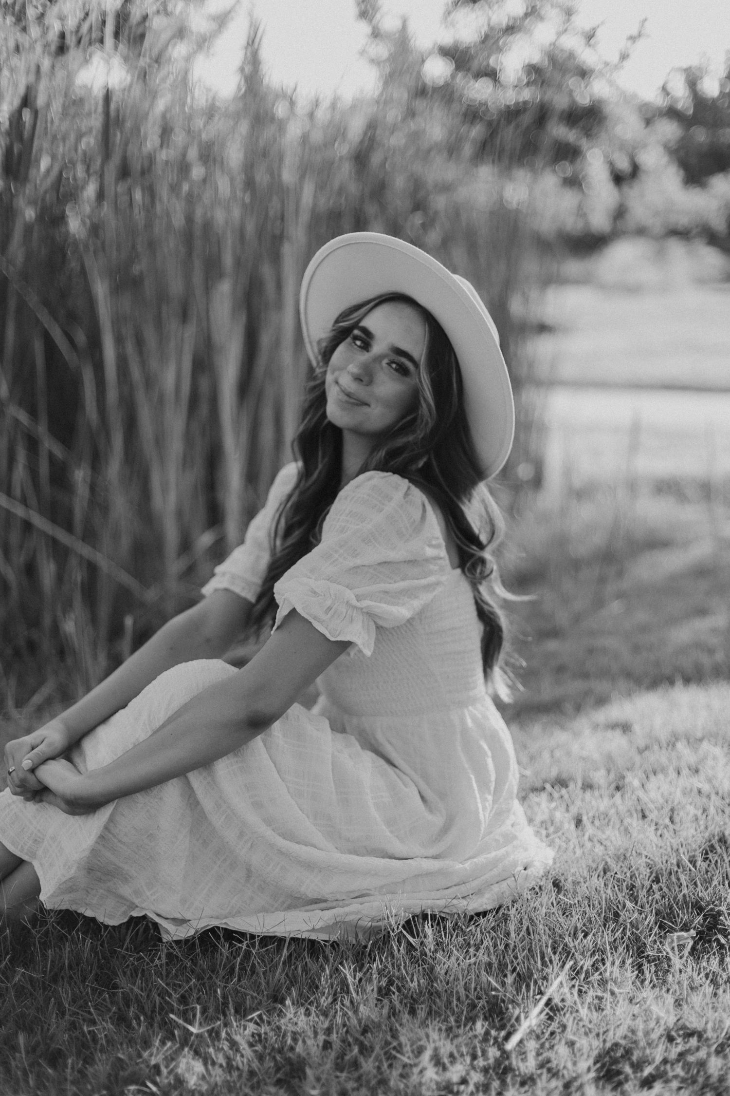 Black and white photo of woman in dress and hat sitting gracefully on grass.