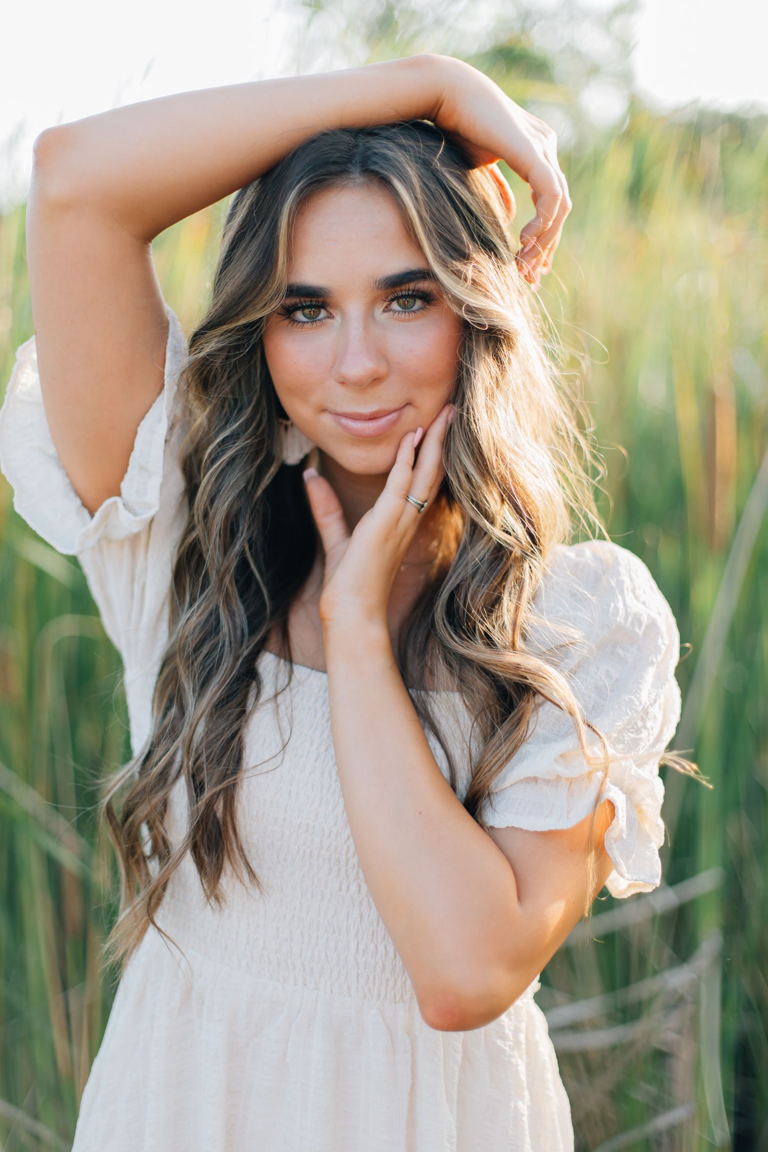Portrait of a woman in a white dress posing in a sunlit field with long wavy hair and natural makeup.