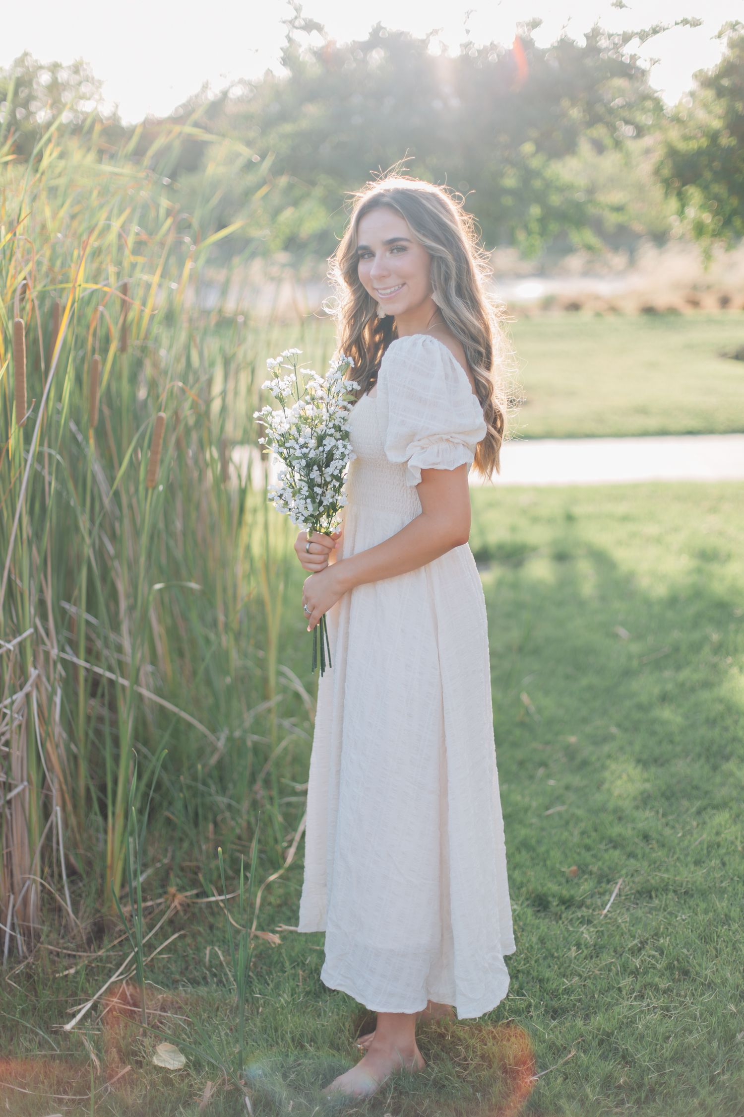 Woman in flowing white dress stands near tall grass in ethereal outdoor setting.