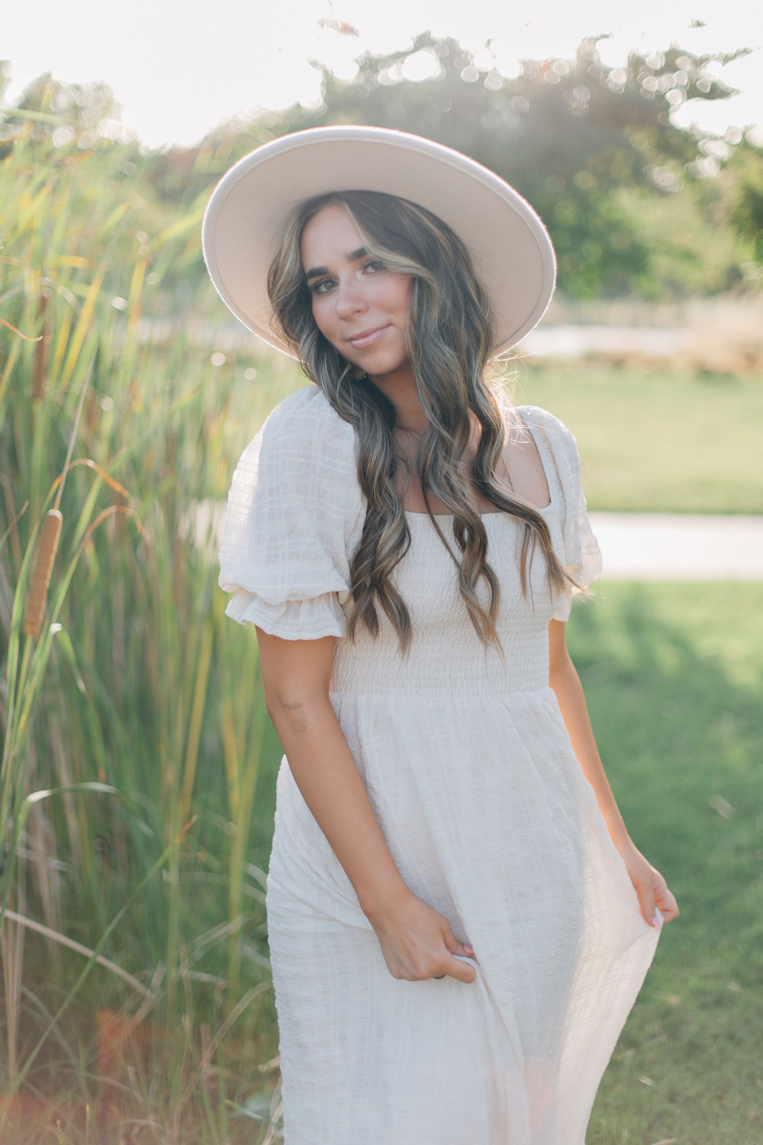 Woman in white dress and wide brim hat poses in front of reeds at sunset.