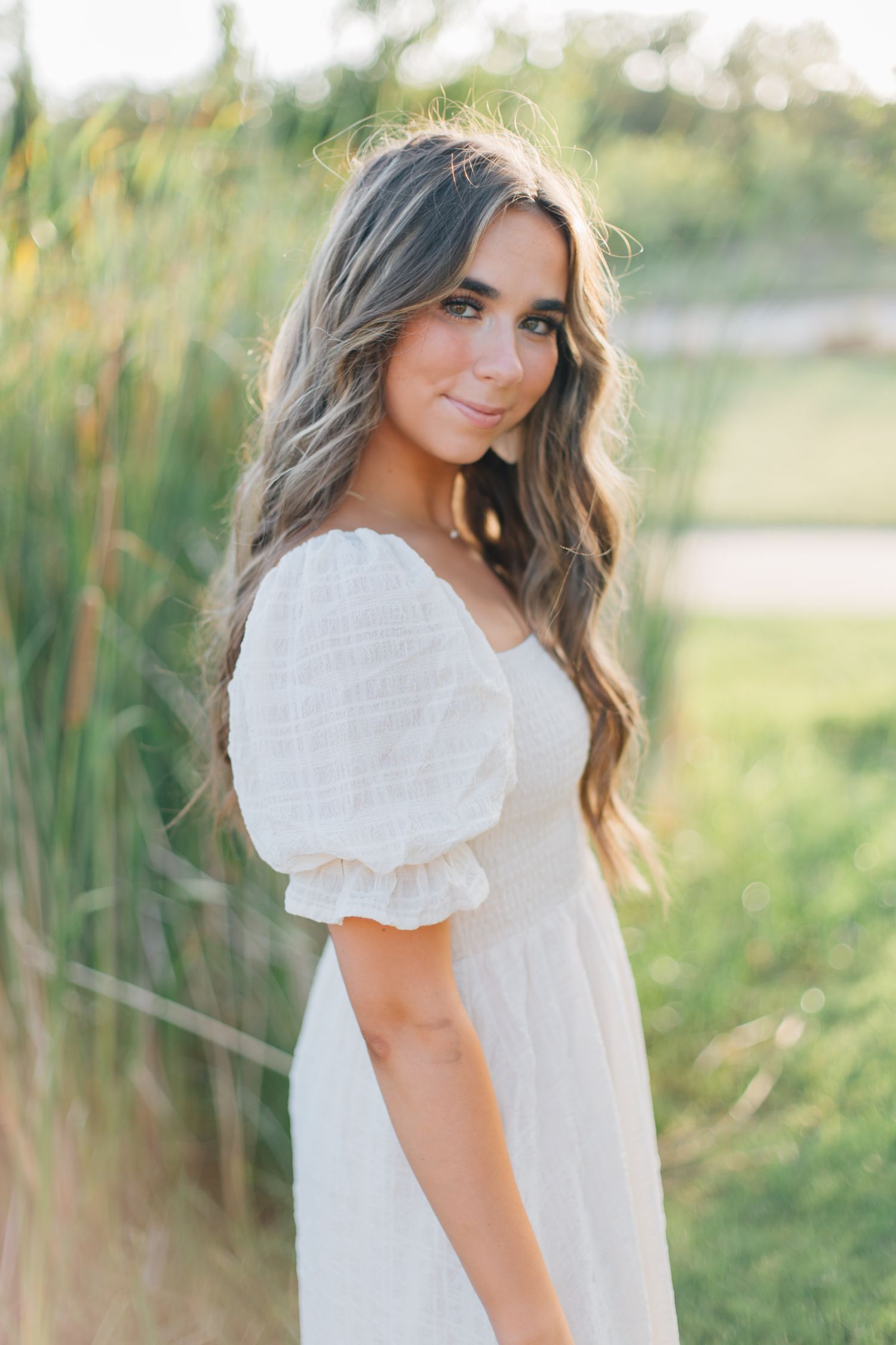 Woman in romantic white dress poses against tall grass in dreamy summer light.
