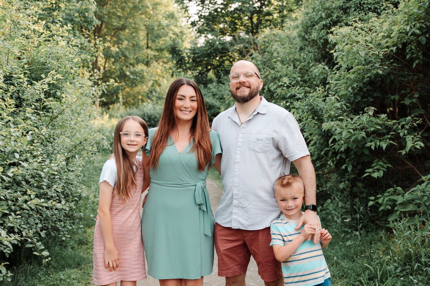 Family of four in coordinated casual outfits poses together in lush green summer setting.