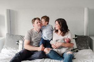 Happy family of four sitting on bed with parents smiling at toddler son while mother holds newborn.