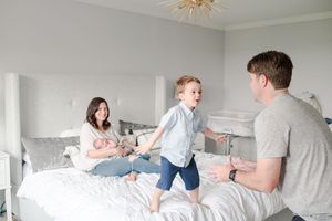 Family playing on a white bed with parents and young boy jumping while mother holds newborn baby.