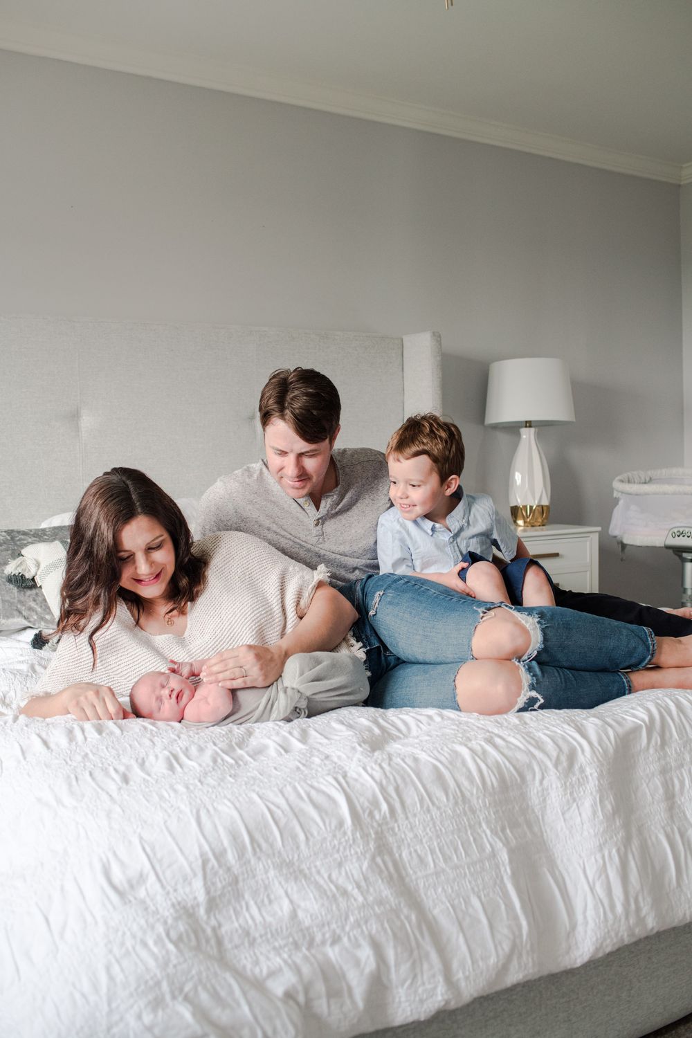 Family of four with newborn baby relaxing together on white bed in bright bedroom.