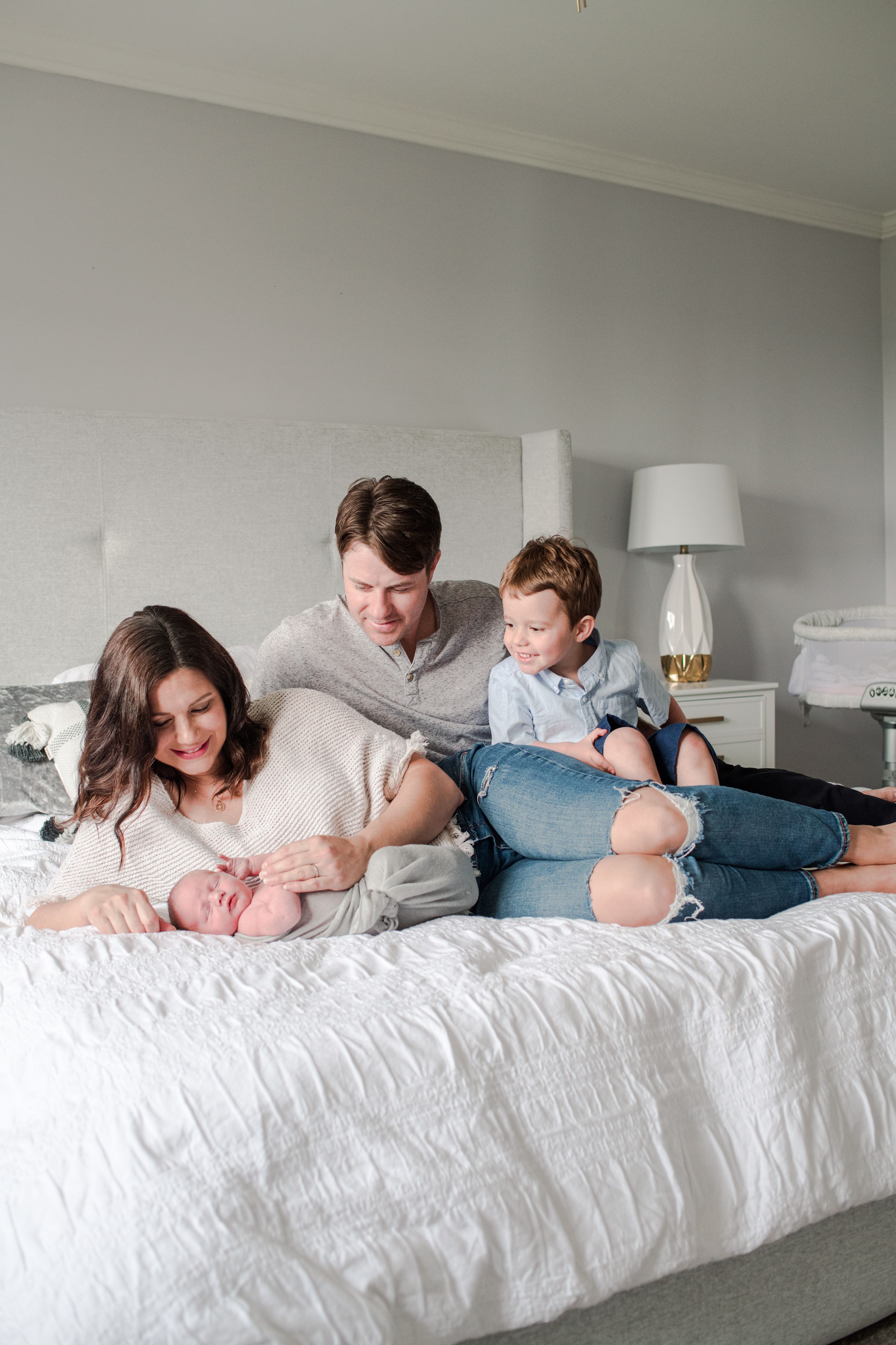 Family of four with newborn baby relaxing together on white bed in bright bedroom.