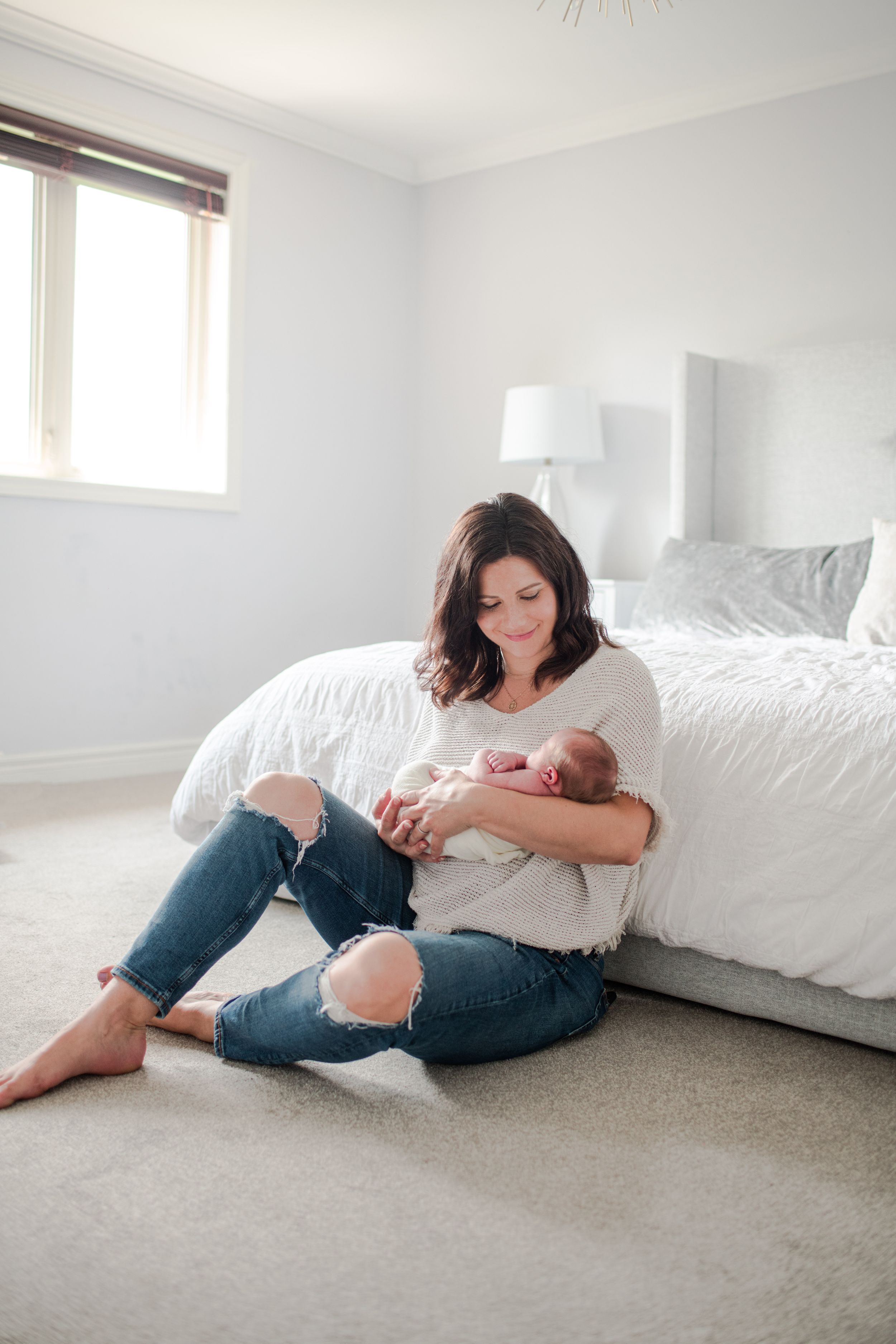 Mother sitting on bedroom floor cradling sleeping newborn baby in her arms tenderly.
