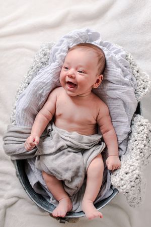 Smiling newborn baby lying in a round basket wrapped in soft gray muslin blankets.