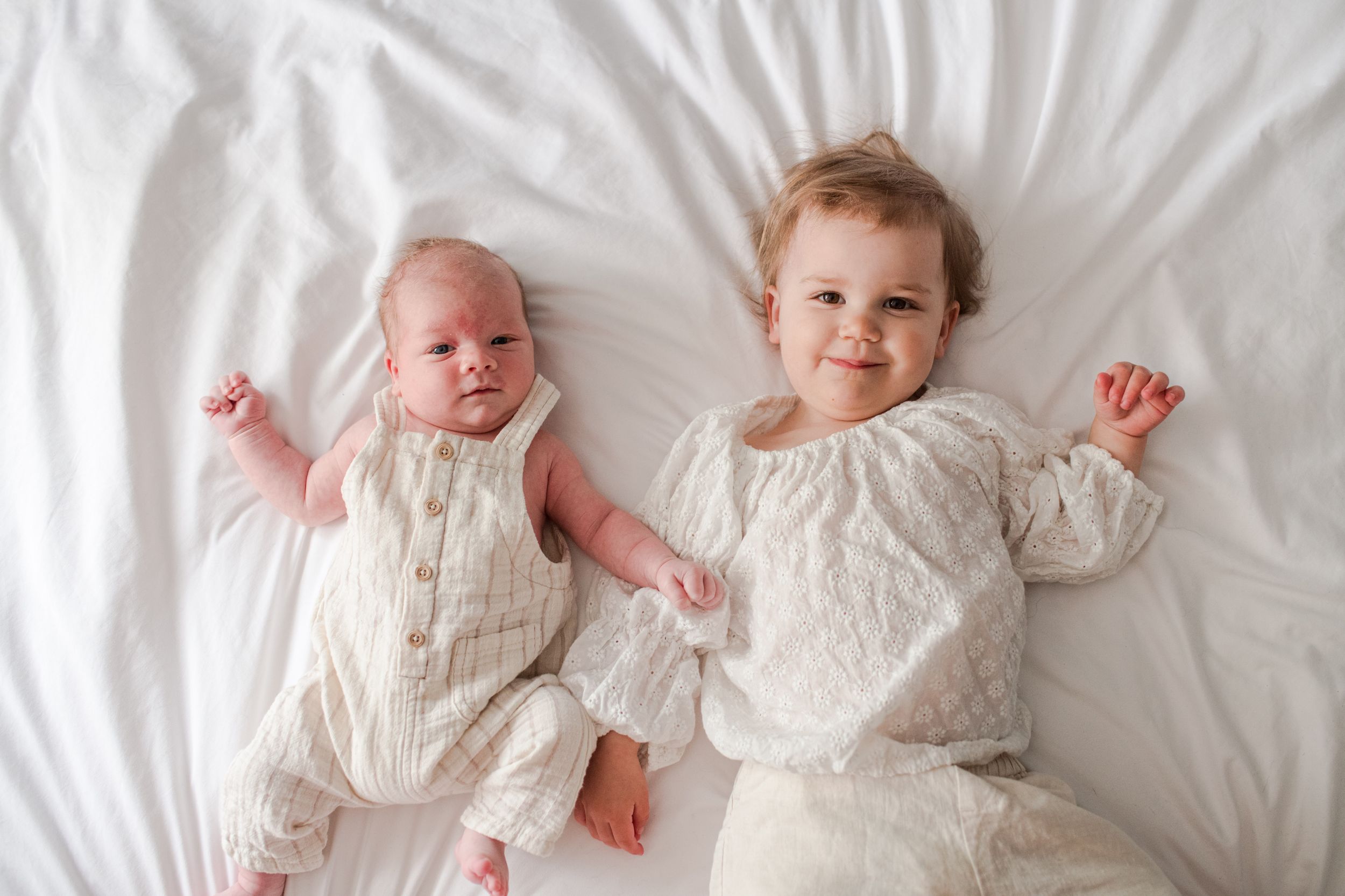 Adorable newborn and toddler sibling lying side by side on white bedding in matching cream outfits.