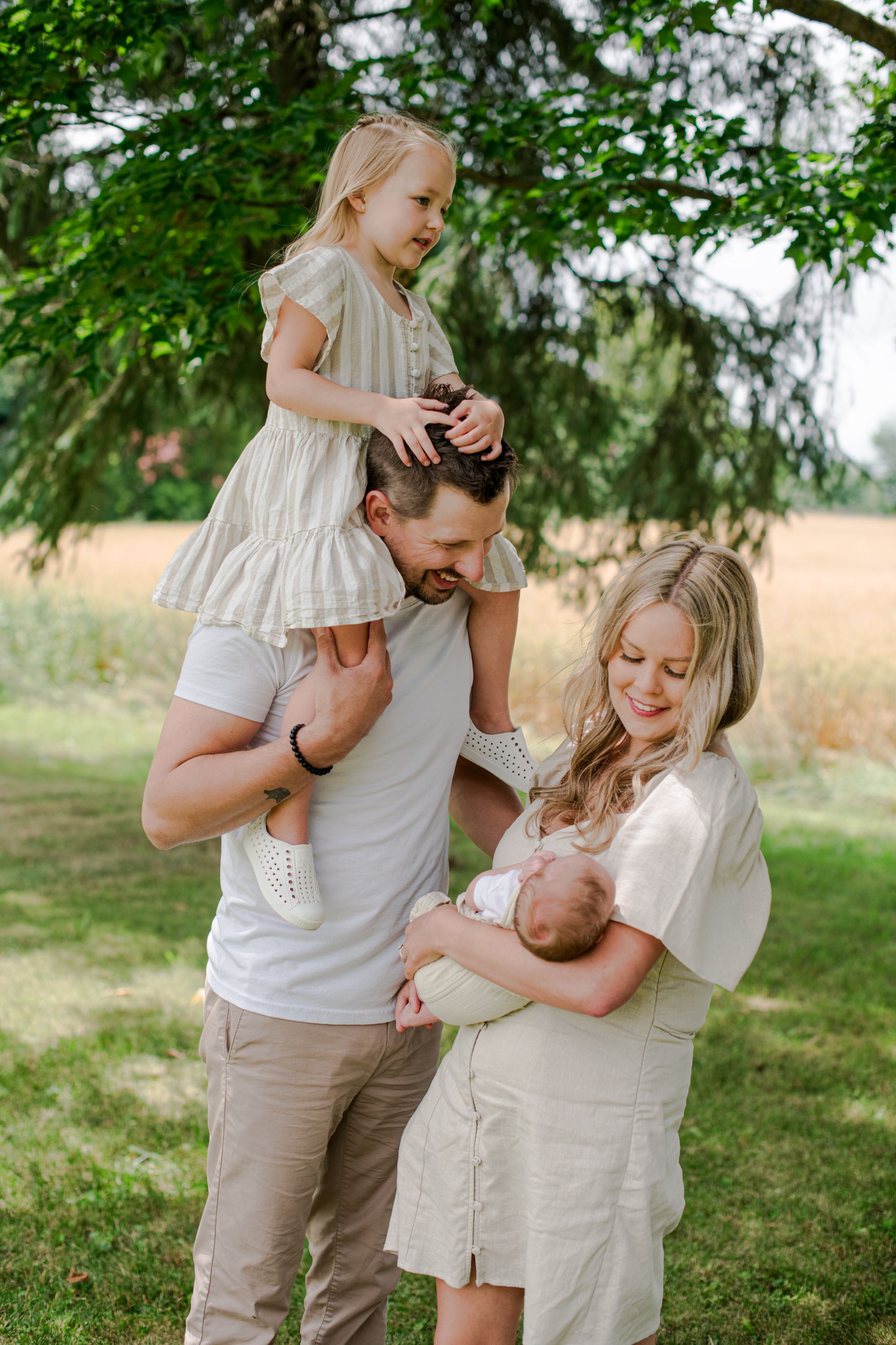 Joyful family in neutral tones outdoors with daughter on fathers shoulders and mother cradling newborn.