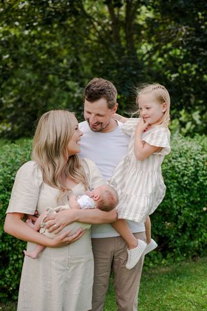 Young family of four standing outdoors with mother holding newborn and father lifting toddler daughter.