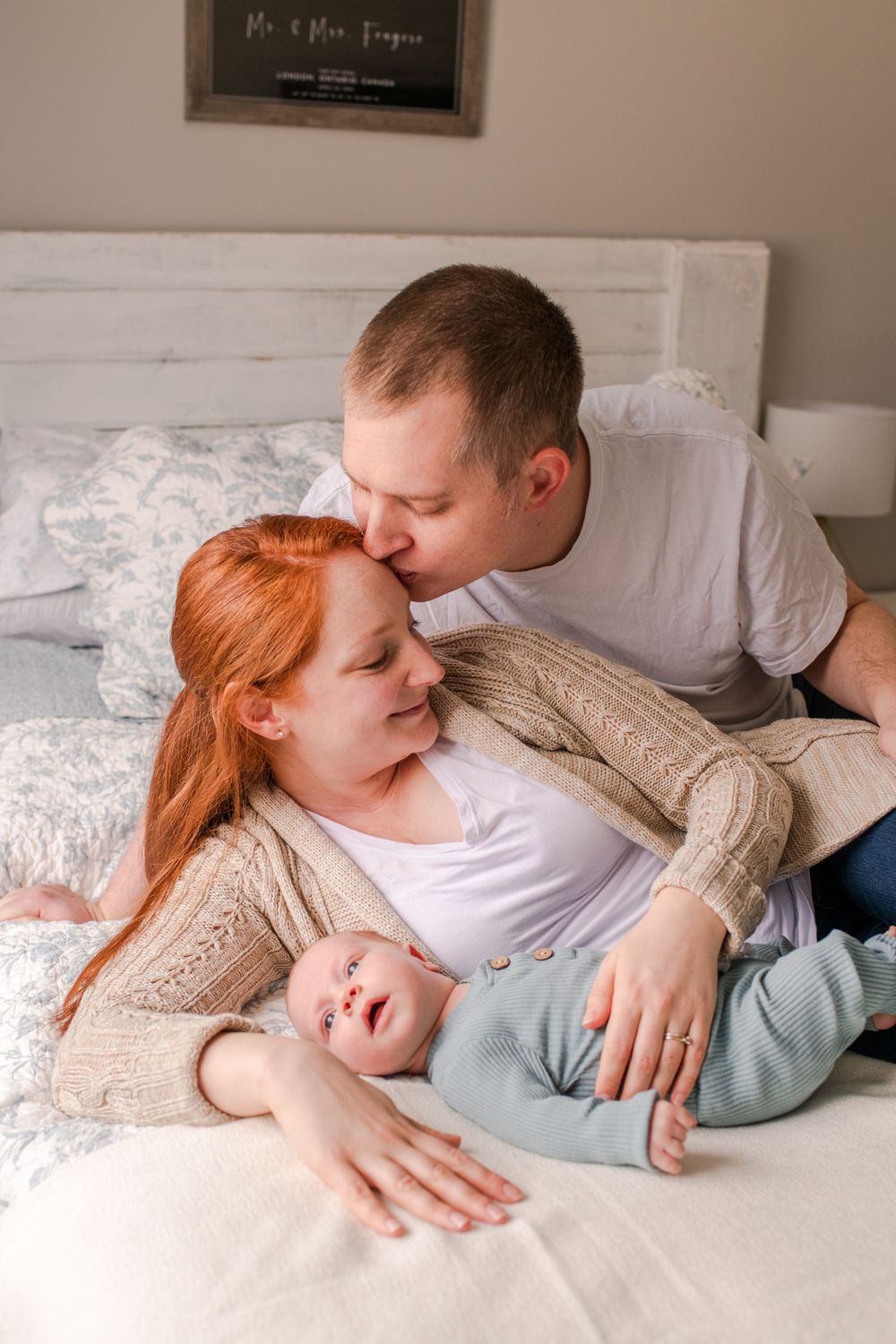 Father kissing mother while she holds newborn baby on white bed in bedroom.