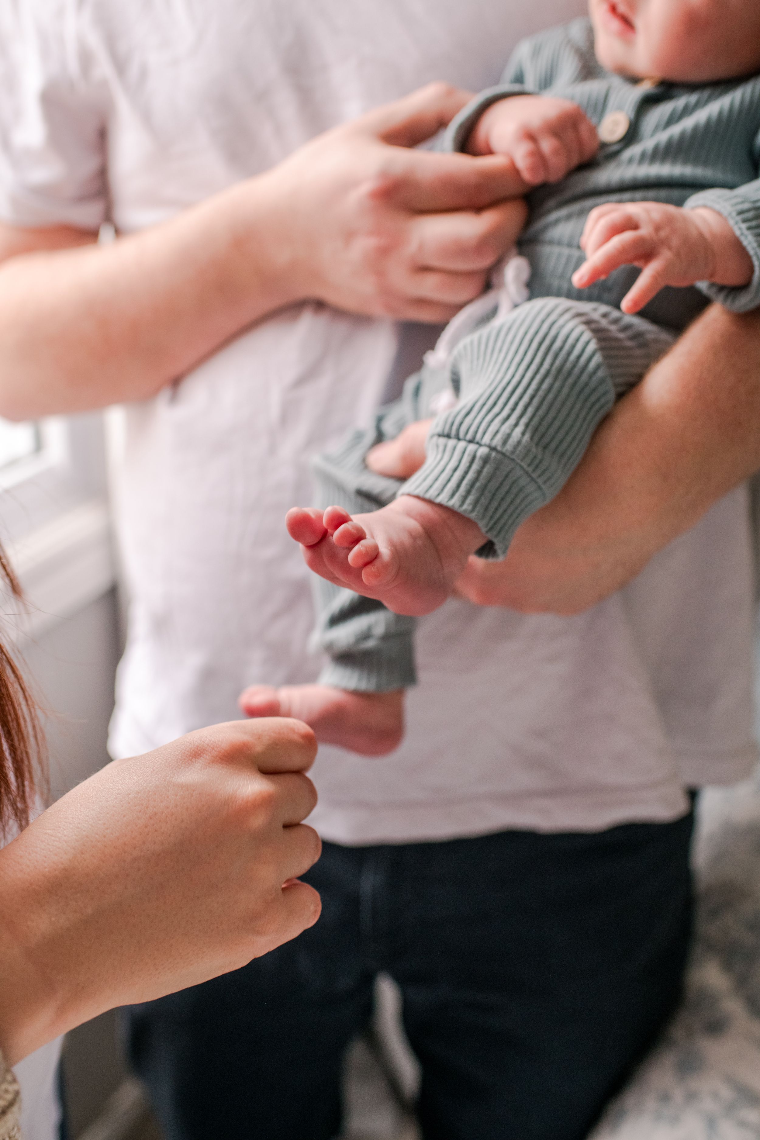 Parents holding newborn baby in sage green outfit, close-up of tiny feet and hands.