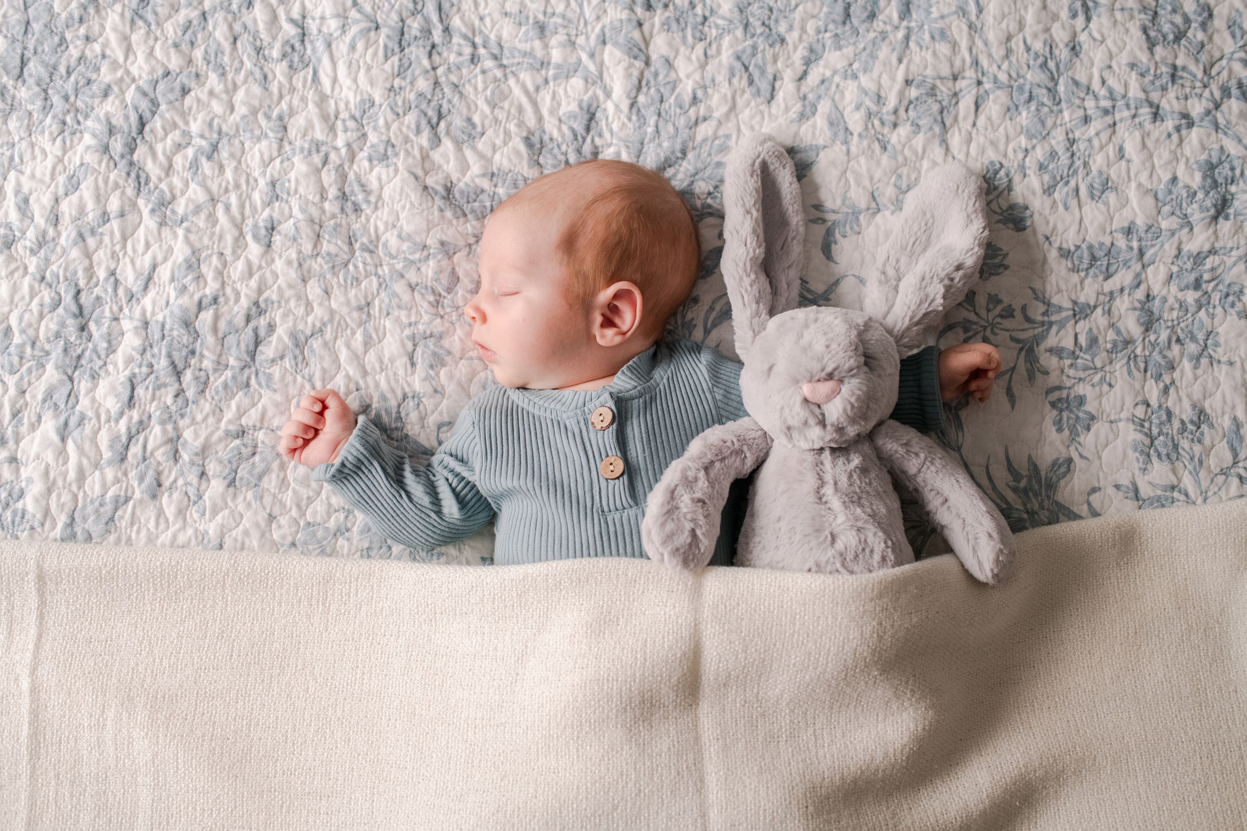 Sleeping newborn baby in sage green outfit beside stuffed bunny on blue textured rug.