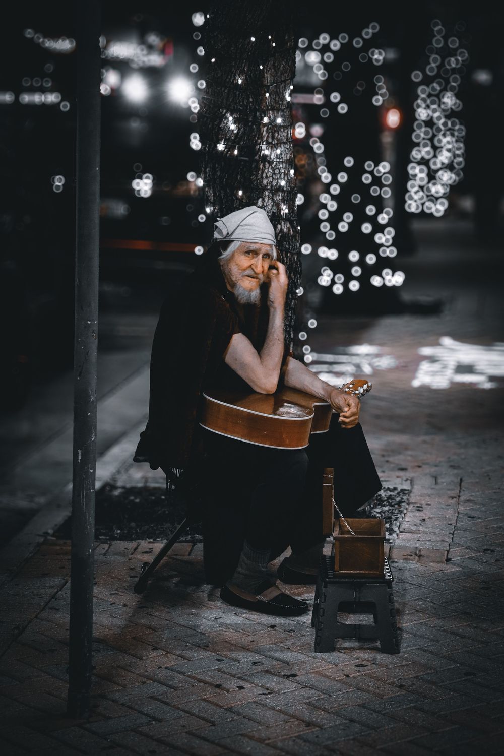Street musician playing acoustic guitar on cobblestone pavement with bokeh lights in background.