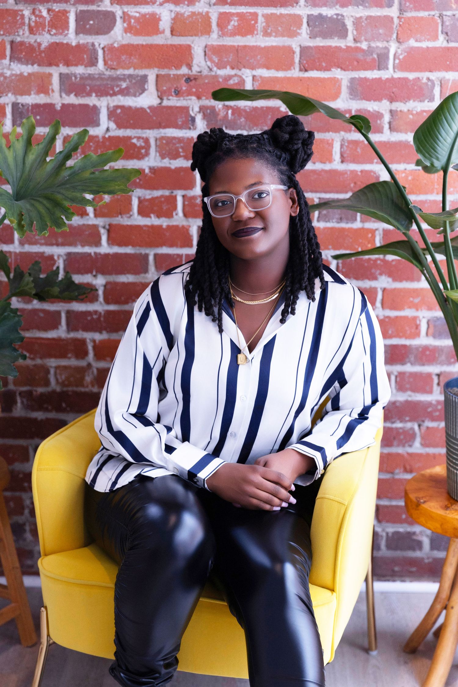 Professional wearing a striped button-down shirt sits in a yellow chair against a brick wall with plants.