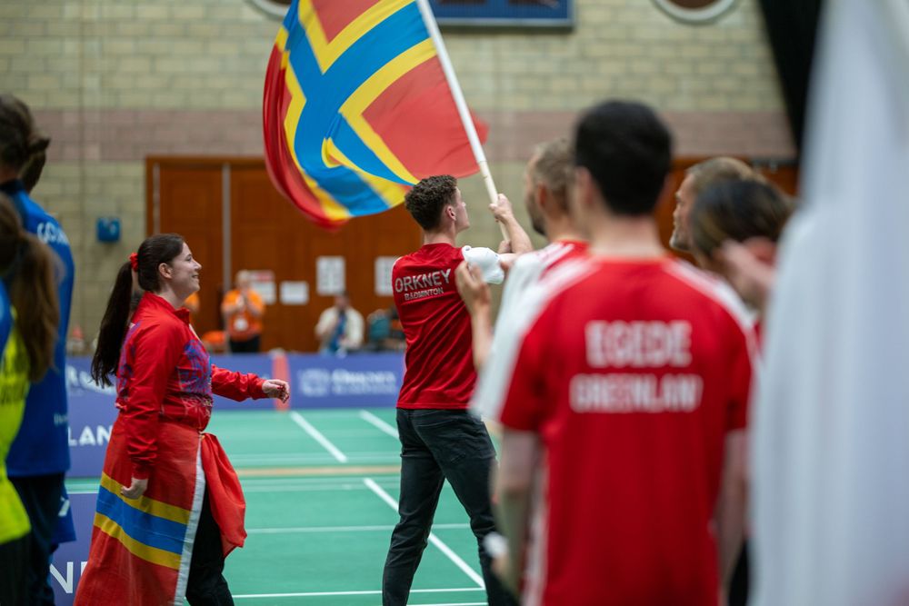 Indoor badminton players compete during a match on green courts with colorful banners and spectators in background.