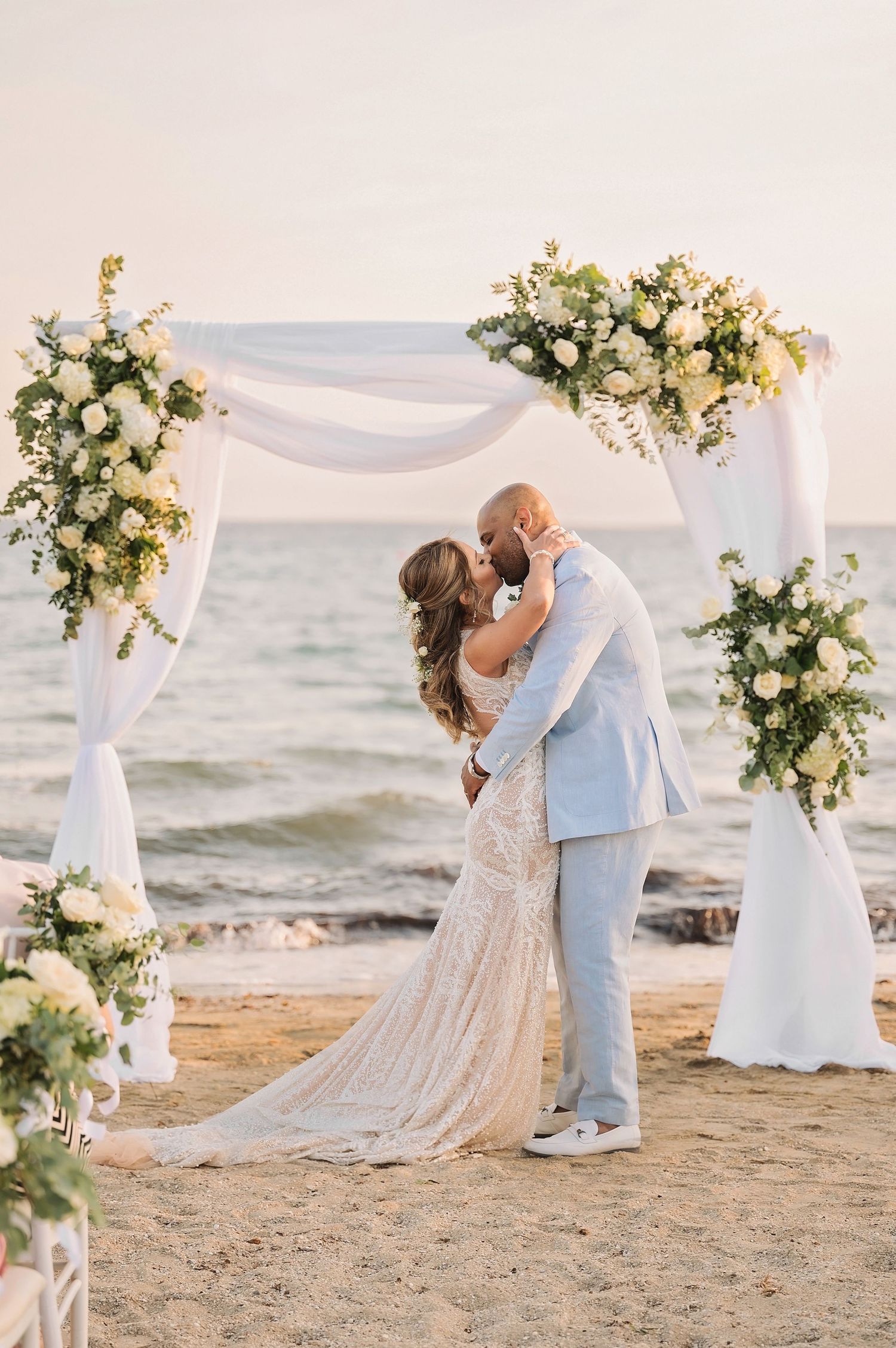 Beach wedding ceremony with couple kissing under floral arch with ocean backdrop.