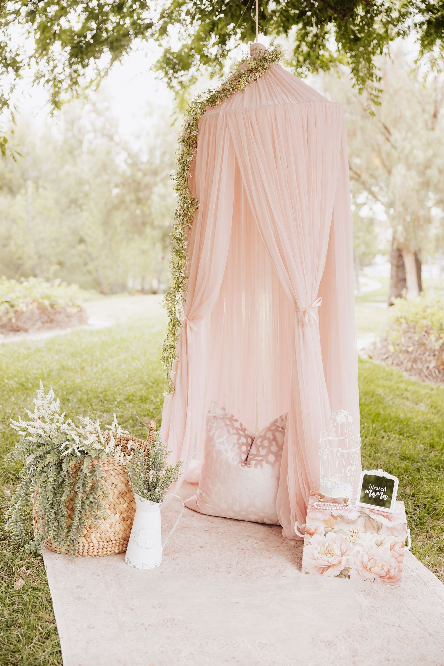 Blush pink canopy tent with floral garland and decorative accessories set up outdoors for a romantic garden party.