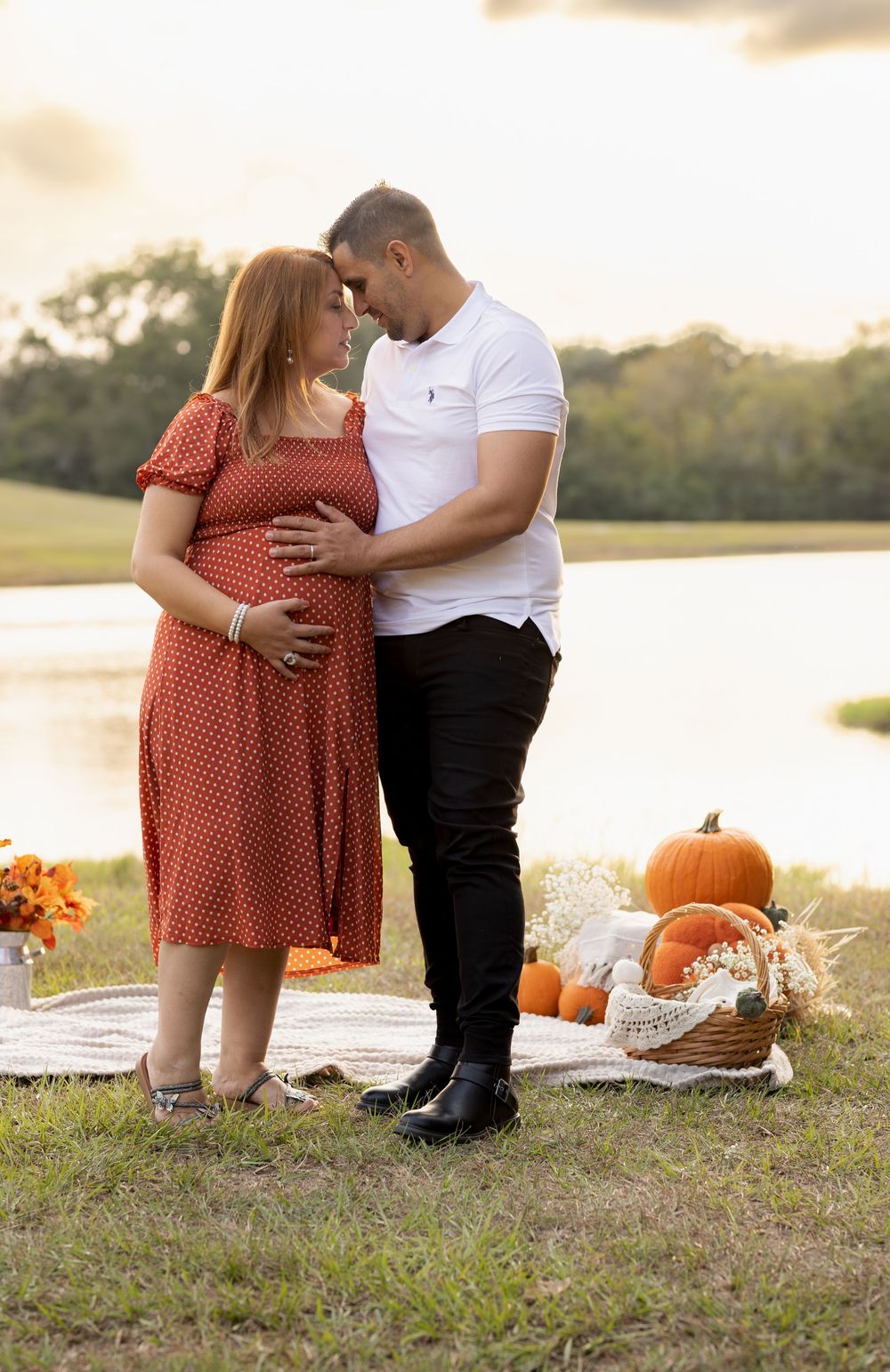 A couple poses with pumpkins and fall decor by a lake during sunset for autumn maternity photos.