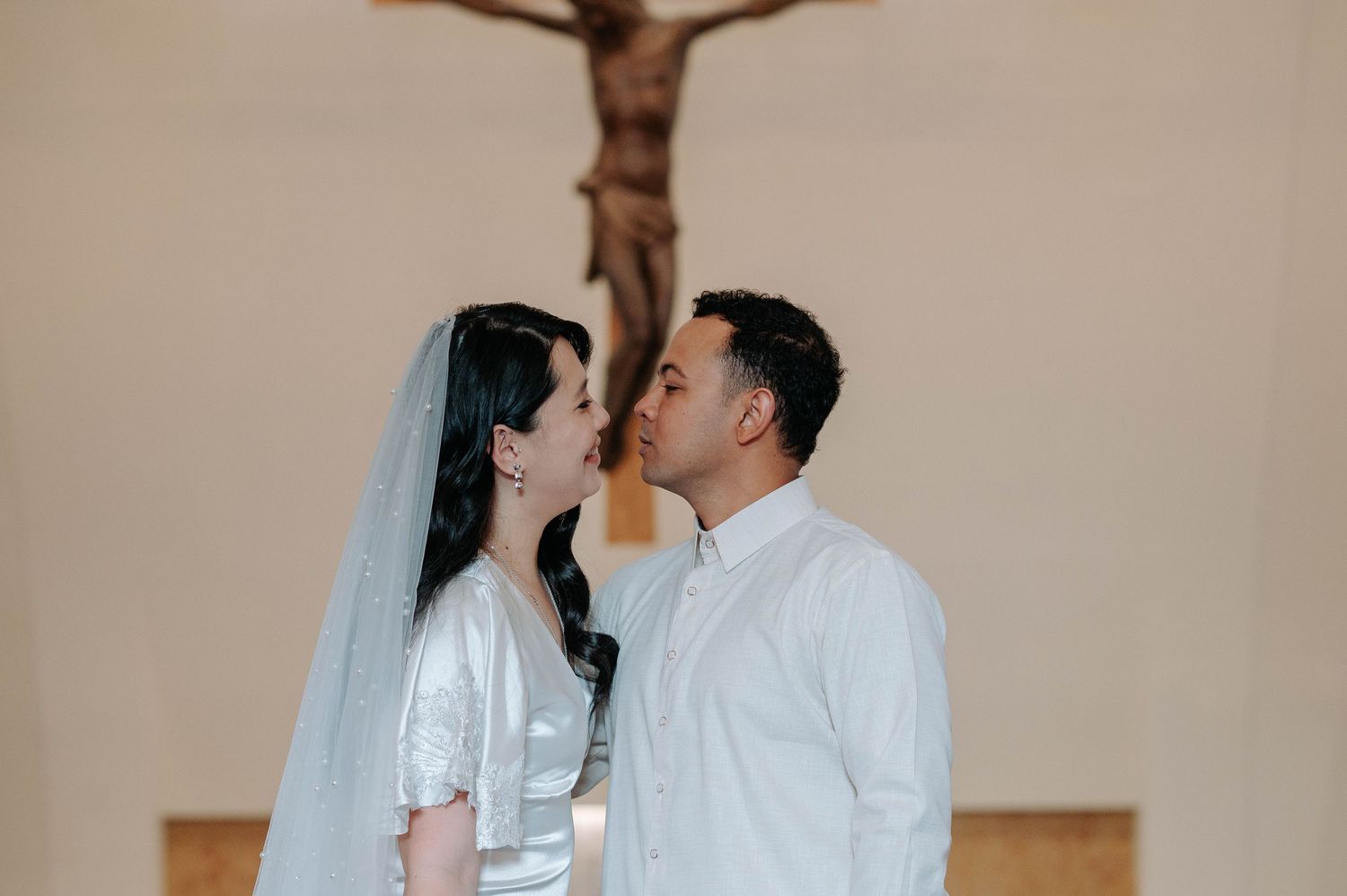 Two people in white attire share an intimate moment in front of a wooden cross at a church ceremony.