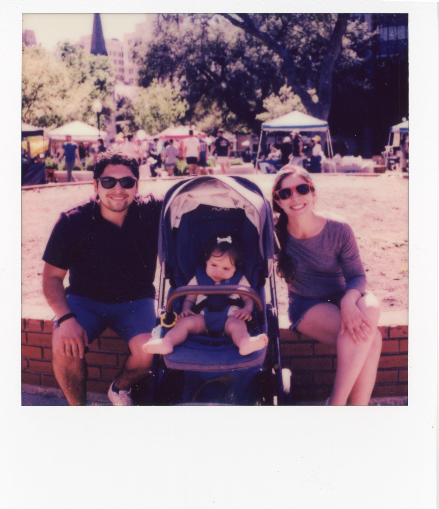 A vintage polaroid photo showing a family sitting on a brick ledge with a stroller at an outdoor event with tents.