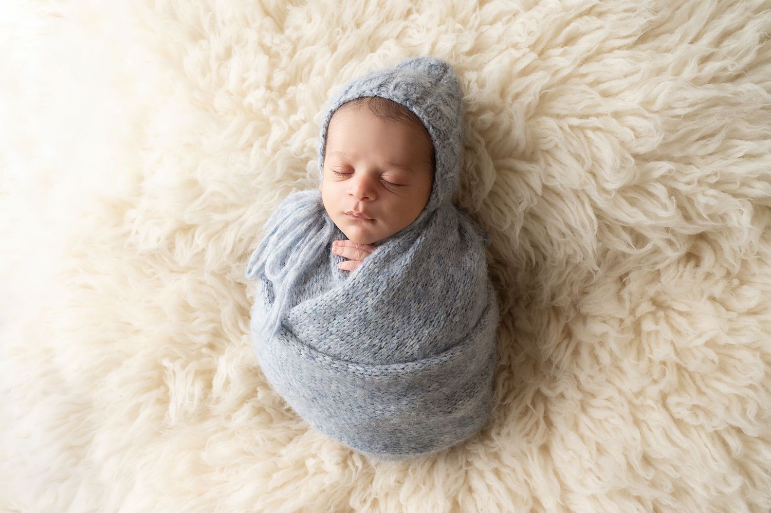 Sleeping newborn baby wrapped in gray swaddle and bonnet lying on plush white fur backdrop.