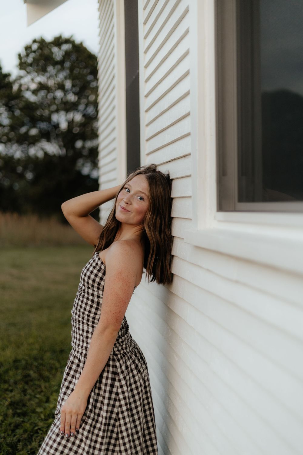 A person in a black and white gingham dress poses against a white house exterior during golden hour.