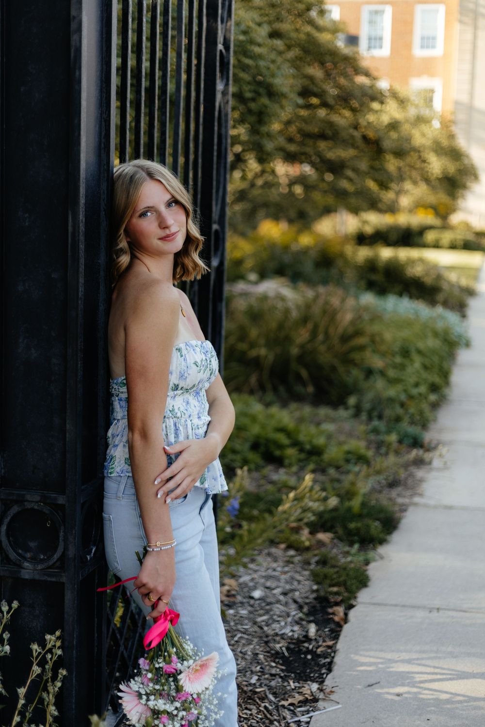 Someone in a white dress poses against an iron fence during golden hour with flowers in hand.