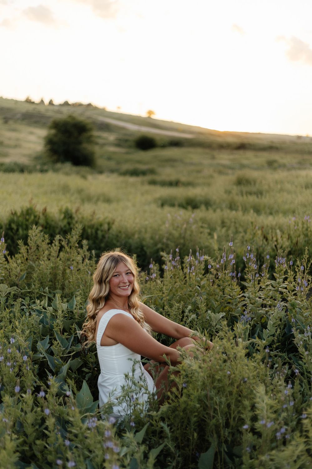 A person in a white dress stands among tall wildflowers in a golden meadow at sunset.