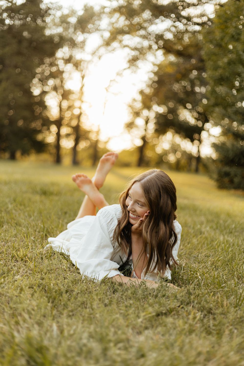 A person in a white dress lies in a grassy field at golden hour, creating a dreamy outdoor portrait.