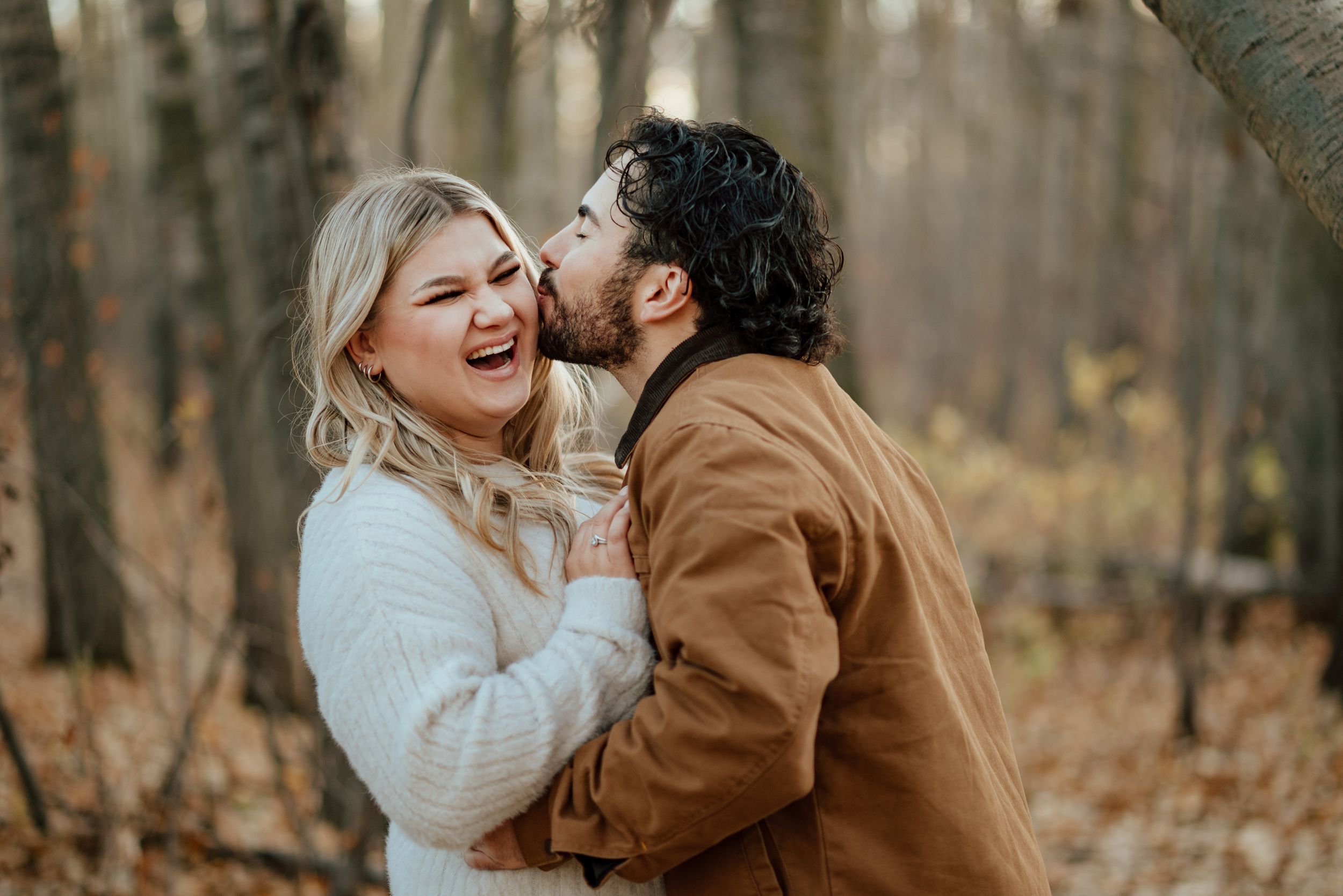 A couple shares joyful moments together in a wooded autumn setting wearing white and brown outfits.