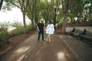 Two people walk hand in hand down a tree-lined pathway surrounded by lush greenery on a summer day.