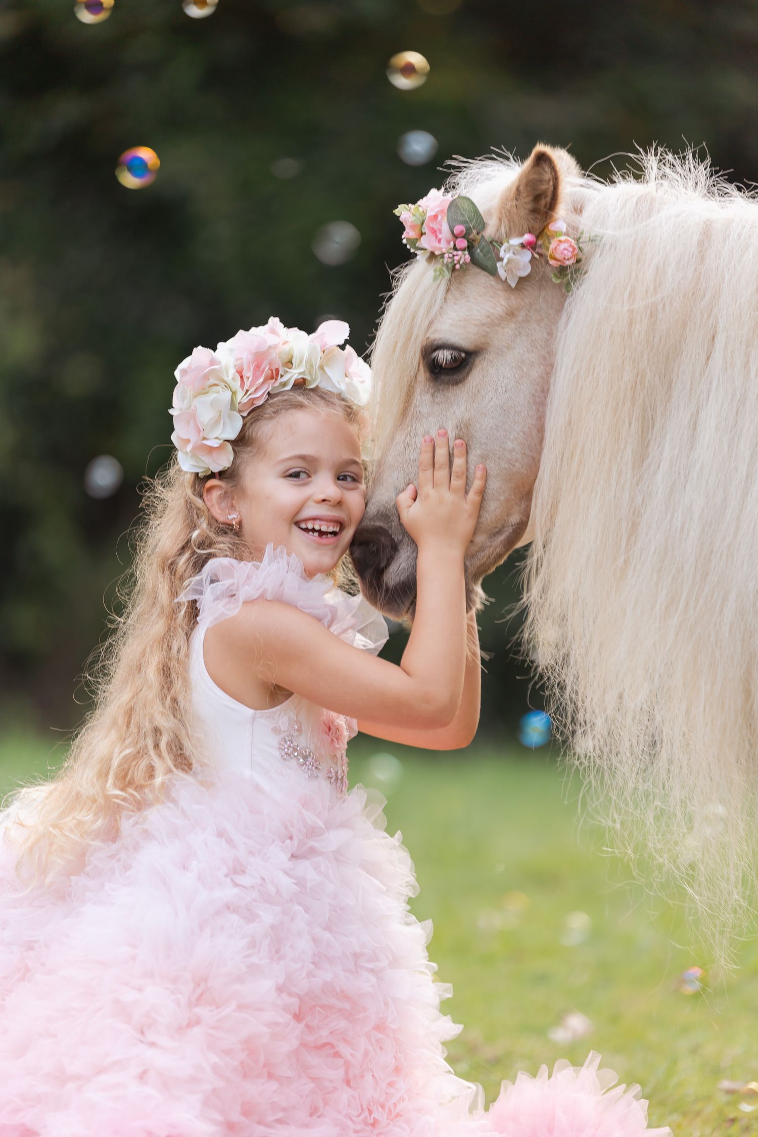 Magical outdoor photoshoot with bubbles, pink tulle dress, and a white horse in a garden.