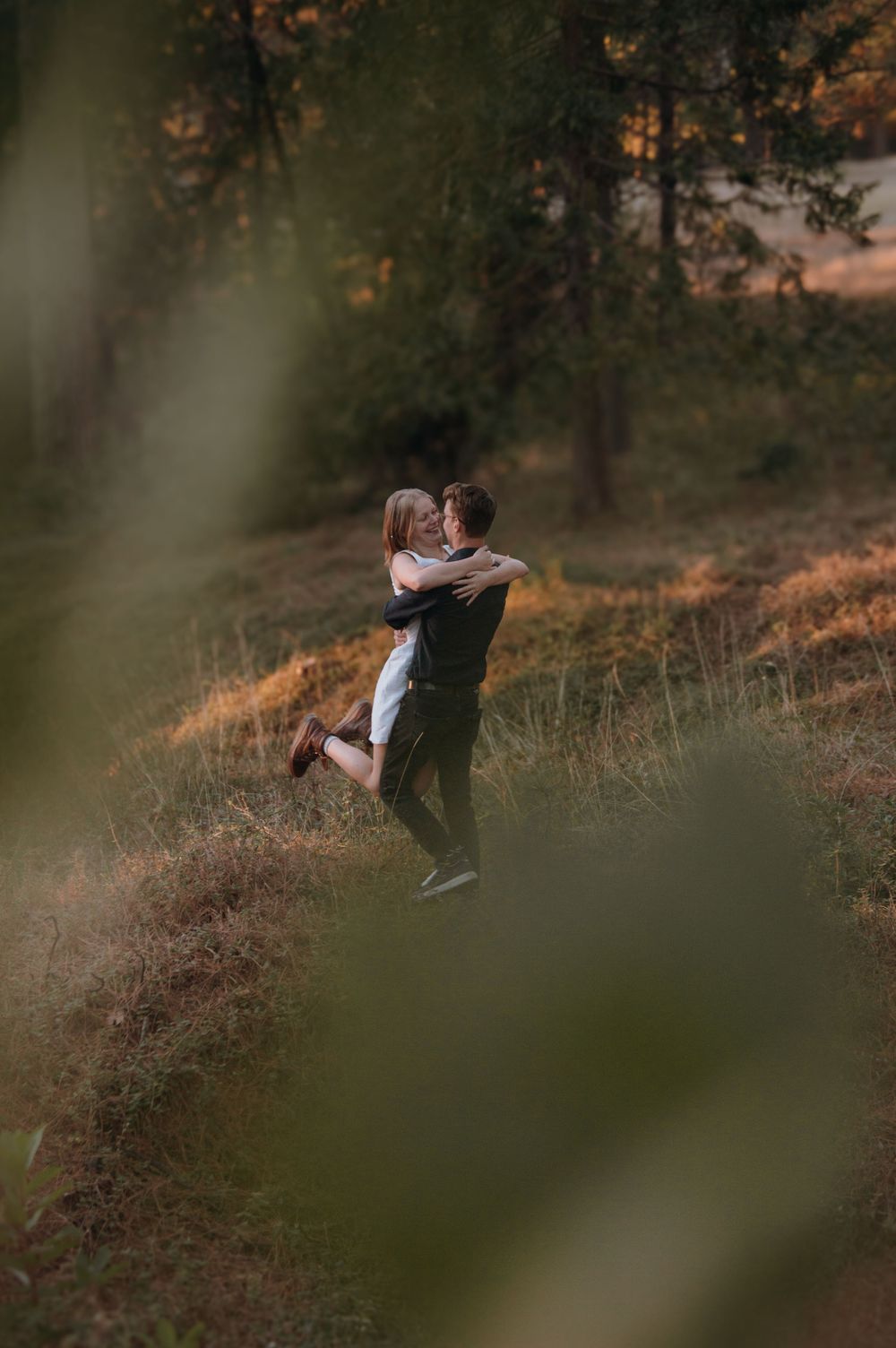 A romantic hillside embrace photographed through blurred foliage at sunset.