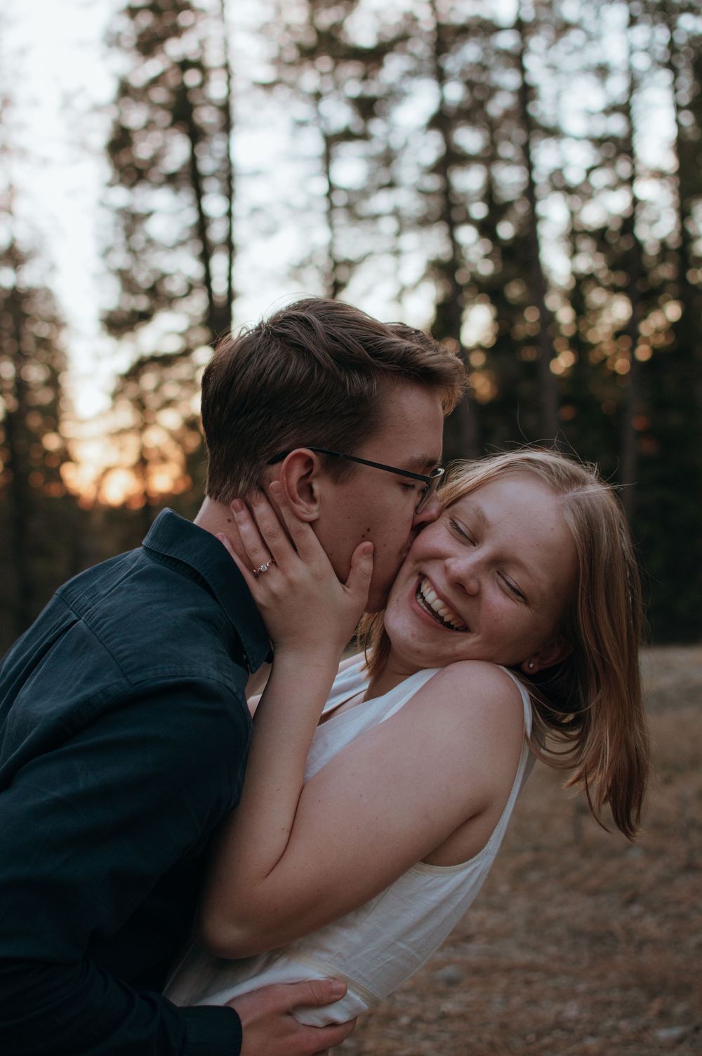 A romantic couple shares an intimate moment laughing together in a forest during golden hour.
