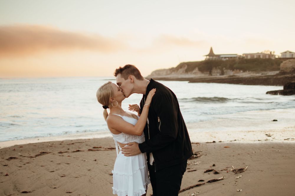 Couple embraces on beach at sunset with coastal cliffs and ocean in background.