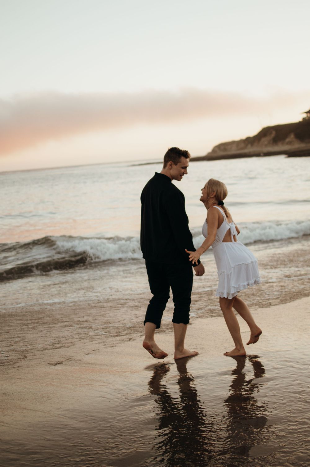 Couple dancing barefoot on wet sand at sunset with ocean waves and cliffs in the background.