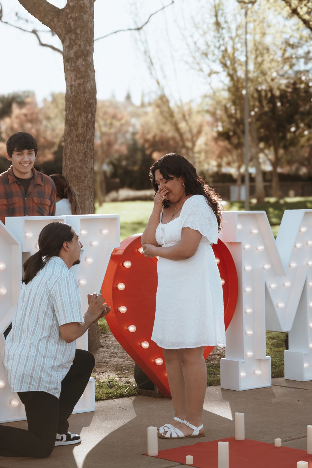 A proposal scene with illuminated letters and a heart decoration in an outdoor setting.