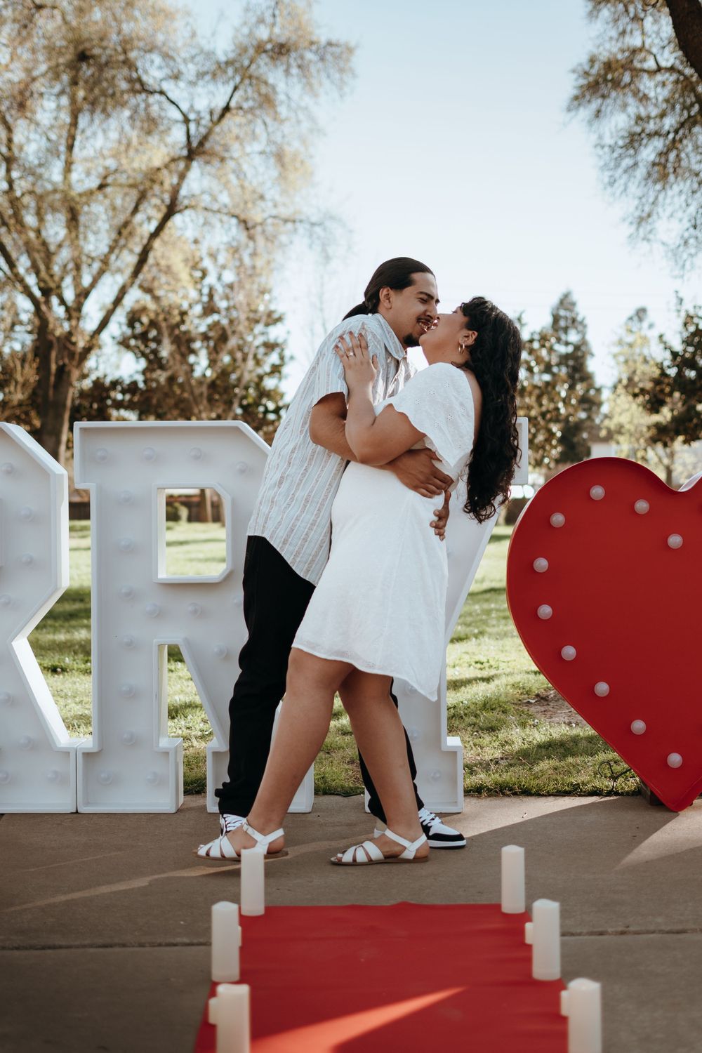 A couple celebrates their engagement in front of decorative light-up letters and a heart prop.