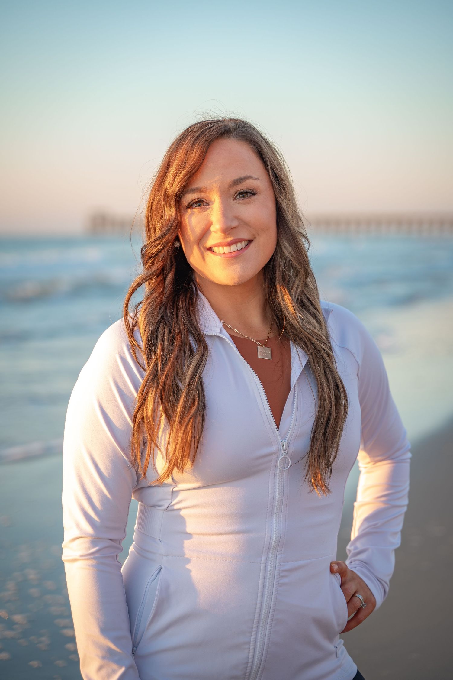 Woman in white zip-up jacket smiling on beach at sunset with ocean pier in background.