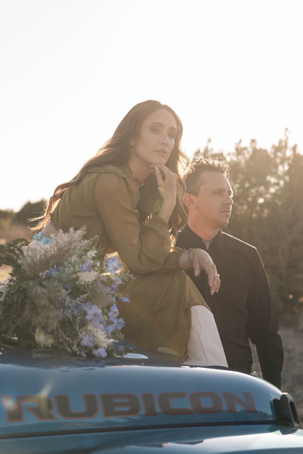 A couple stands beside a Jeep Rubicon at sunset, with flowers and warm backlighting creating a romantic atmosphere.