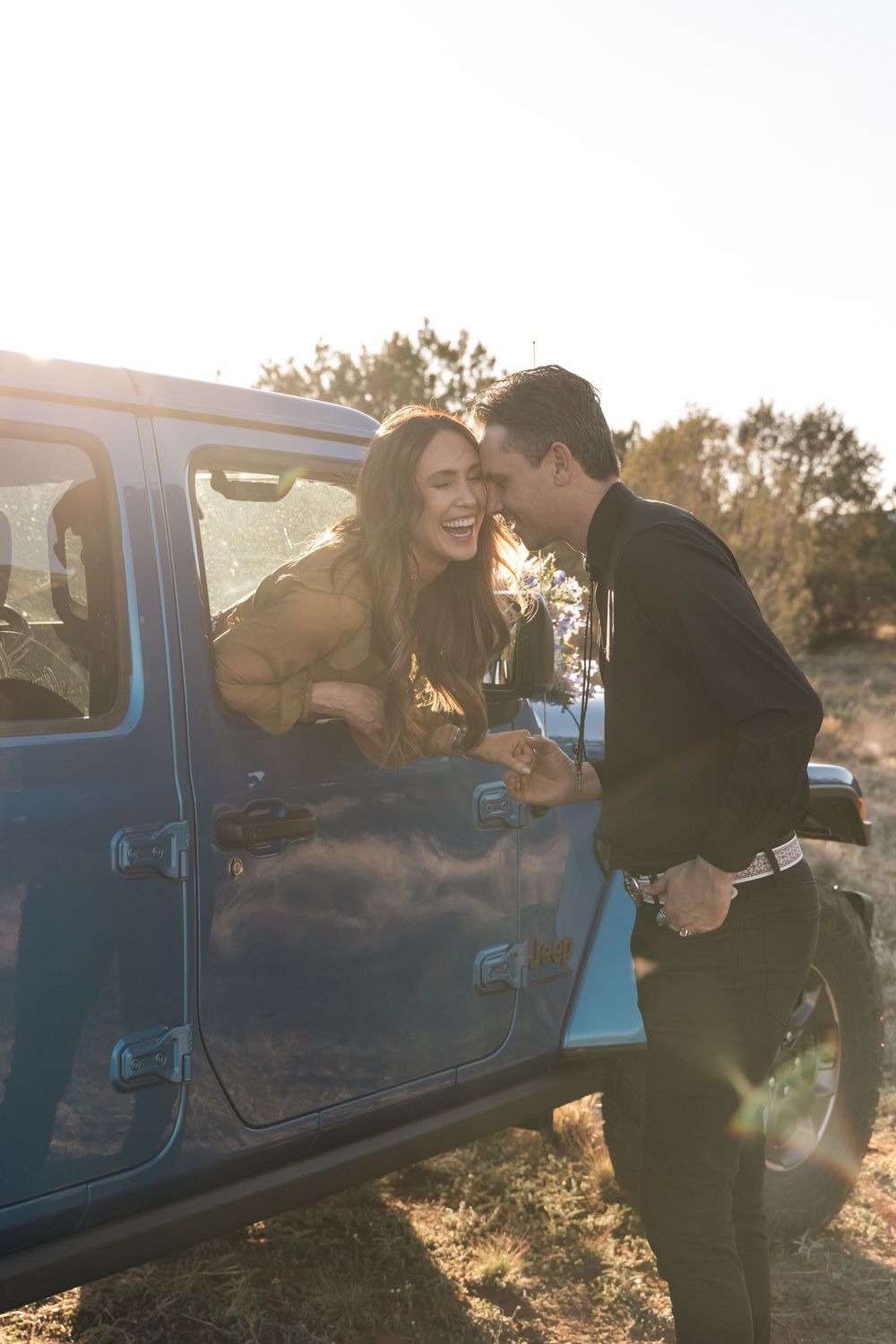 A couple shares a joyful moment together by a blue Jeep during golden hour sunset.