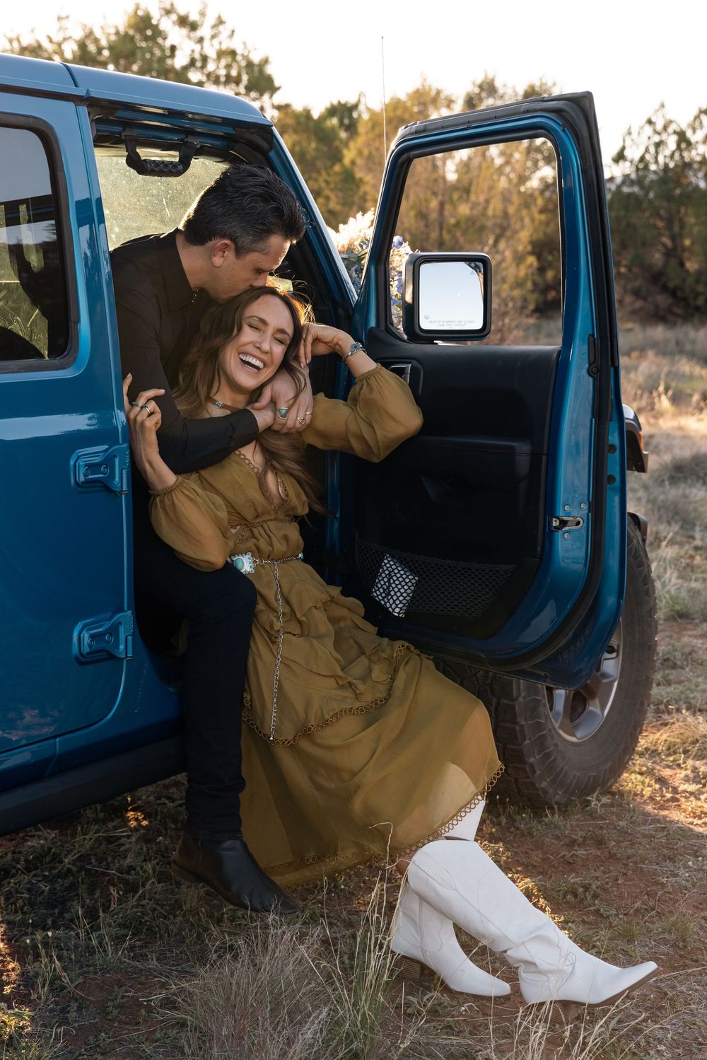 Two people share a joyful moment while sitting in a blue truck during golden hour in a natural outdoor setting.