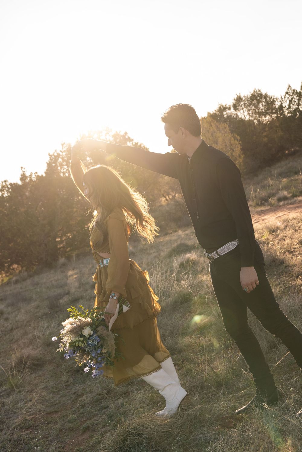 Couple walking and dancing together on a hillside at sunset with sunflare, woman in yellow dress holding flowers.