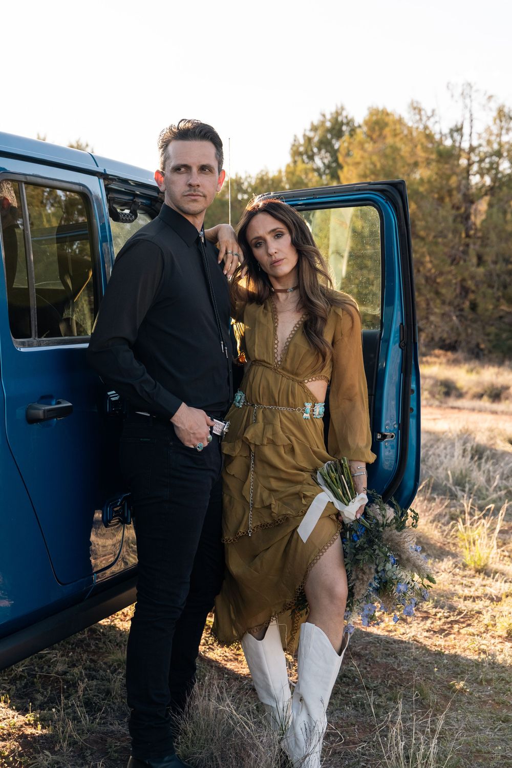 Couple poses beside vintage blue vehicle in desert setting with woman wearing brown dress and white boots.
