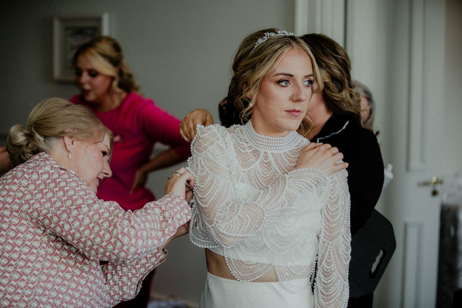 Two people adjusting a delicate lace wedding dress with high neckline and crop top design.